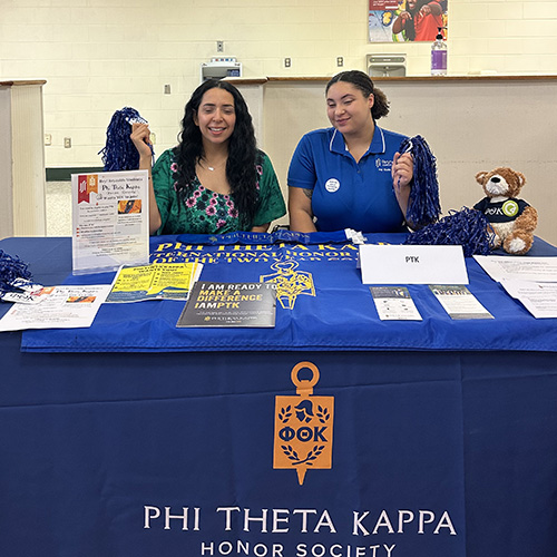  Alyssa and Fab, two Reynolds students, sit at a table with P.T.K. information and giveaways smiling at the camera and holing pom poms in one hand