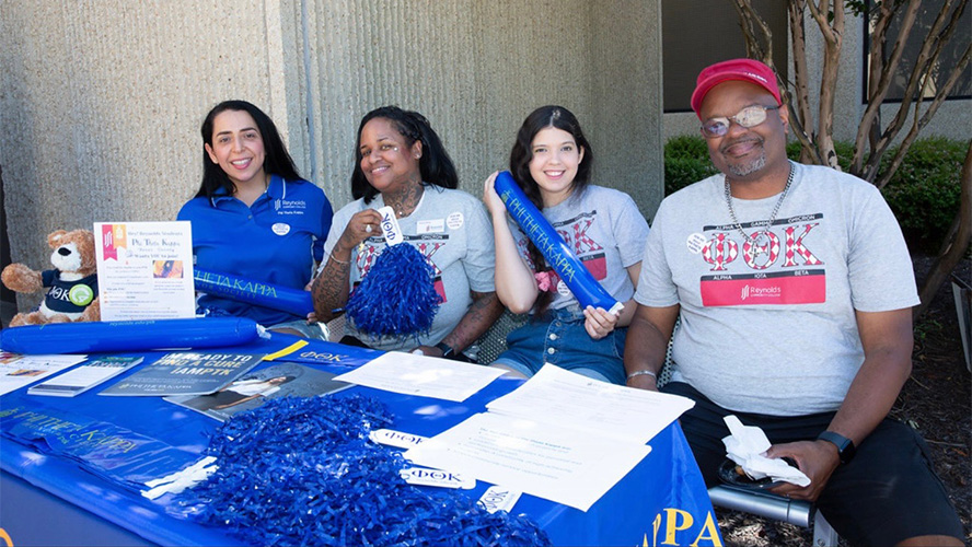 four students sit a table with P.T.K. information and giveaways smiling at the camera