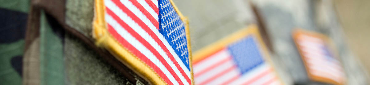 Close-up of an American flag patch with gold trim on military camouflage fabric, with additional flag patches visible but out of focus in the background